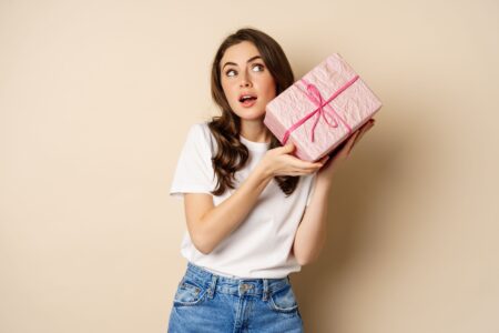jpeg optimizer celebration holidays concept happy young girl looking intrigued shaking box with gift guessing whats inside wrapped present standing beige background